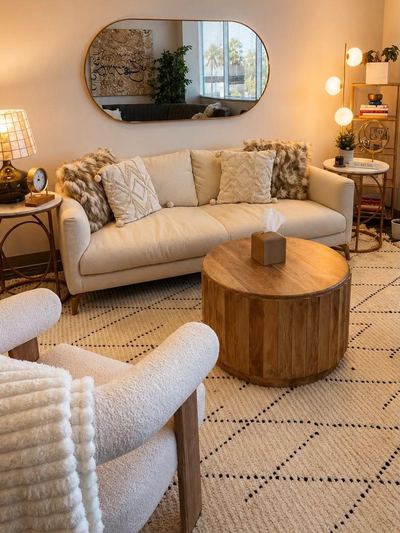 A cream-toned individual therapy room with a soft sofa, boucle armchair, and warm lamplight